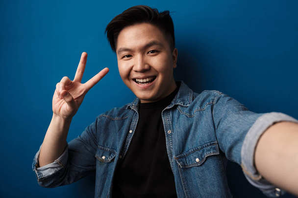 Image of laughing handsome asian man gesturing peace sign and taking selfie photo isolated over blue background - Φωτογραφία, εικόνα