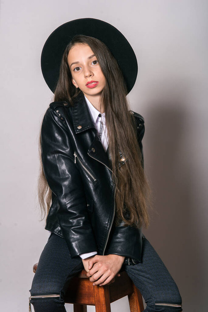 Egy tinédzser lány divatos bőrdzsekiben és egyenletes kalapban, fehér háttérrel. Tinédzser divat. Utcai divat. Szépség és divat - Fotó, kép