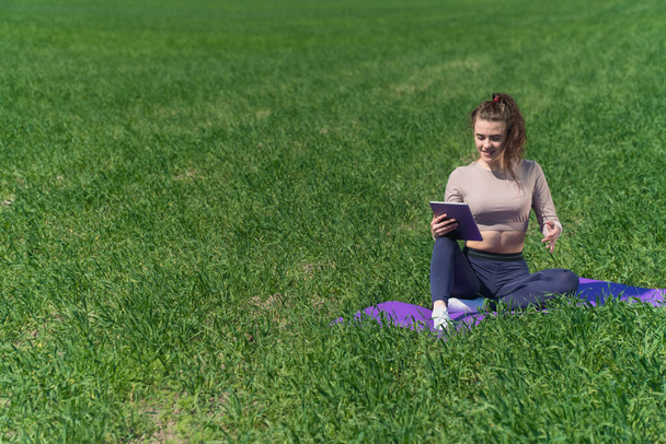 A young girl looks into a digital tablet against the backdrop of nature in a field. Stylish fitness coach female looks into smartphone sitting on yoga mat - Valokuva, kuva