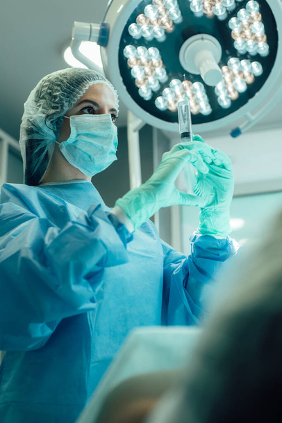 Σοβαρή γιατρός στέκεται κάτω από μια χειρουργική λάμπα δίπλα στον ασθενή της και κοιτάζοντας τη σύριγγα στα χέρια της - Φωτογραφία, εικόνα