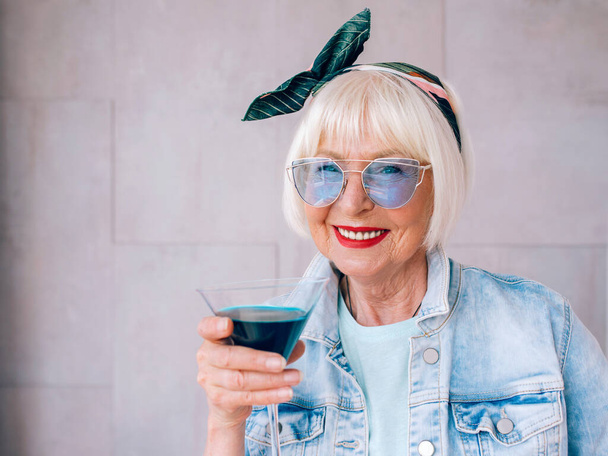 mulher (velha) elegante sênior com cabelo grisalho e em óculos azuis e casaco de ganga segurando vidro com coquetel azul. Álcool, relaxamento, férias, conceito de aposentadoria
 - Foto, Imagem