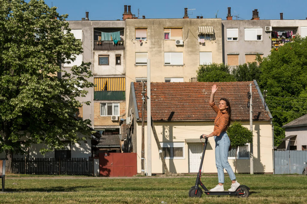 Young beautiful woman rides an electric scooter through an urban settlement, while waving in the direction of a photographer. - Фото, зображення
