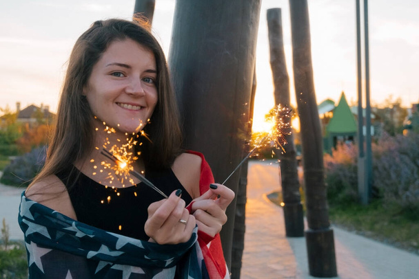 Щаслива усміхнена молода жінка з іскрами та портретом прапора США на заході сонця в парку - Фото, зображення