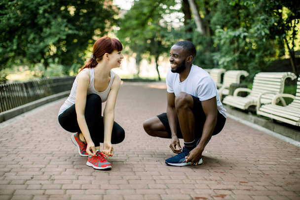 Smiling young multiethnic active couple, African handsome man and pretty Caucasian woman, tying trainer shoe laces before jogging in park, looking each other. Sport and running concept - Foto, immagini