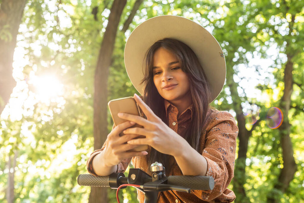 Młoda piękna kobieta w kapeluszu stojąca obok skutera elektrycznego podczas pisania na telefonie. W tle dominują drzewa i zielone kolory. - Zdjęcie, obraz