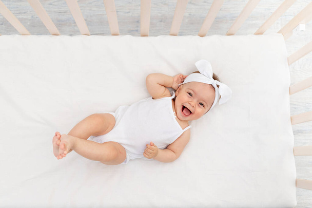 bébé fille 6 mois se trouve dans un berceau dans la crèche avec des vêtements blancs sur le dos et rit, regarde la caméra, le matin de bébé, concept de produits pour bébé - Photo, image