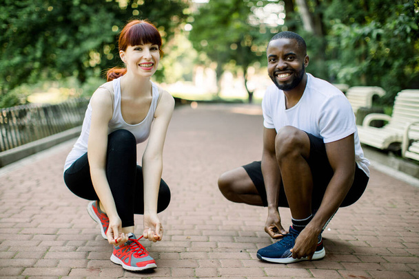 Runners couple, African guy and Caucasian girl, tying running shoes laces at park, smiling and looking at camera. Healthy lifestyle, jogging motivation, happy healthy people - Photo, image
