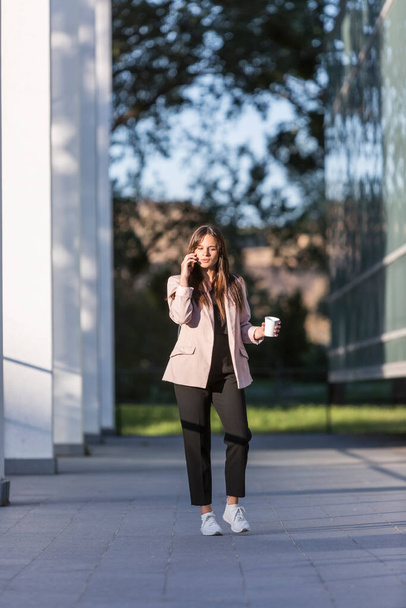 Una giovane bella donna cammina con una tazza di caffè mentre parla sul suo cellulare. Sorride, si forma con l'architettura moderna sullo sfondo. - Foto, immagini