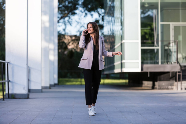 A young beautiful woman is walking while talking on her cell phone. She is smiling, business-trained with modern architecture in the background. - Photo, image