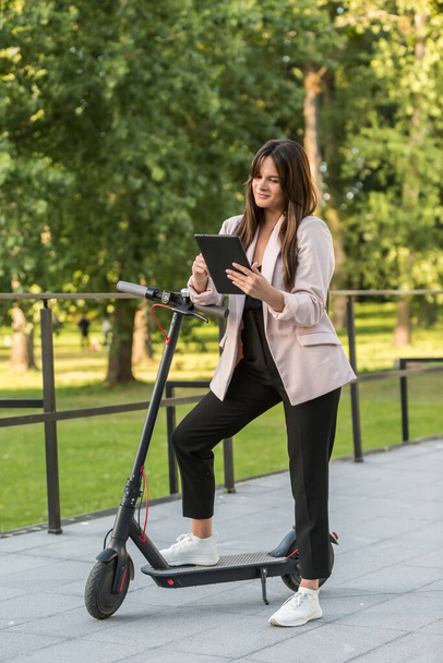 A young beautiful woman is standing next to her electric scooter, with one foot on a standing platform. She is smiling, business-trained, with trees in the background. - Foto, Imagem
