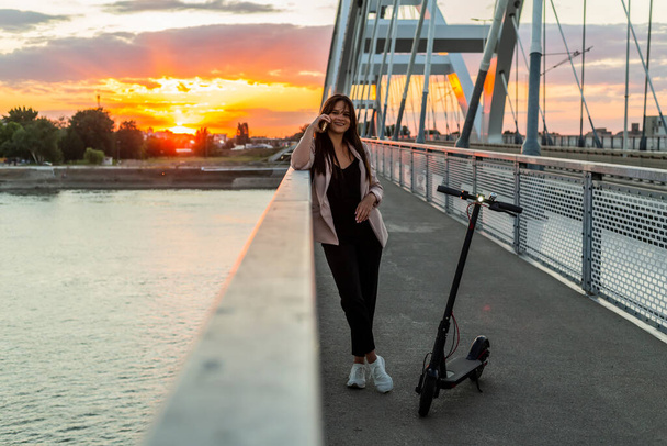 A young smiling woman is standing by the bridge fence while talking on her phone. An electric scooter is parked next to her, while the buildings, sunset and a bridge fence predominate in the background - Fotoğraf, Görsel