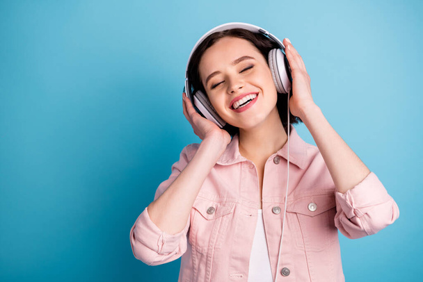 Photo of beautiful cheerful lady short black hairdo hold arms on cool modern technology headphones eyes closed enjoy youth music song wear casual pink jacket isolated blue color background - Fotó, kép