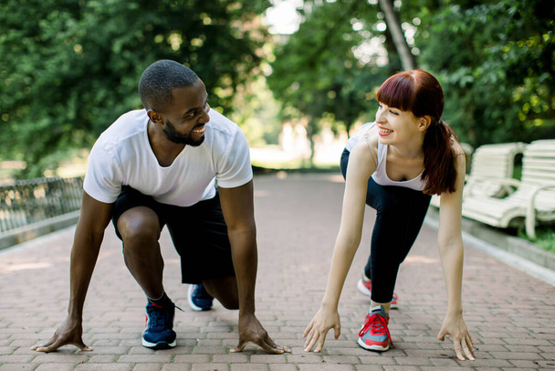 Multiethnic couple in sport clothes on starting line preparing to run in the park, smiling and looking each other. Healthy fit and sportive couple starting position ready for running - Fotó, kép