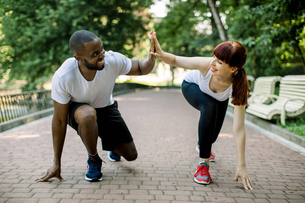 Young fitness couple of African bearded man and attractive Caucasian woman, ready to start running at the park, giving high five each other and smiling. Jogging together outdoors - Fotó, kép