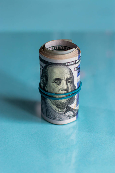 Close up photograph of a rolled up wad of cash sitting on edge with a blue rubber band wrapping across Benjamin Franklin's face on a hundred dollar bill on a white background with copy space. - Photo, Image