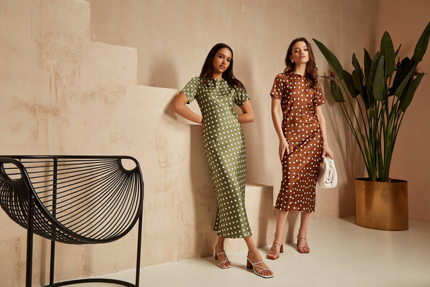 Two fashion model brunette hair wear green  brown dots silk dress sandals  accessory clothes date party walk interior journey summer collection plant flowerpot stairs beautiful woman tan skin friends. - 写真・画像