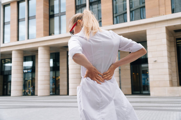 A woman in white medical clothing is holding on to her back. The girl's lumbar pain. The concept of a doctor with a sore back - Fotoğraf, Görsel