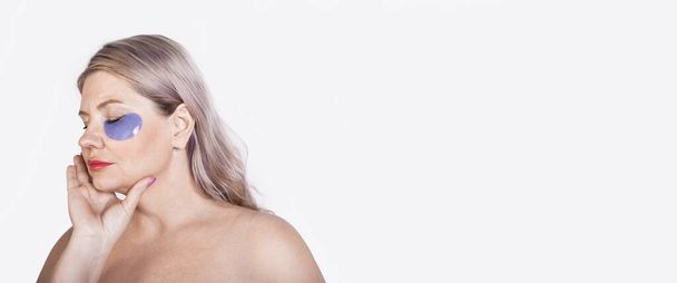 Gealterte kaukasische Frau mit blonden Haaren und Hydrogel-Augenklappen posiert mit nackten Schultern auf einer weißen Studiowand mit freiem Raum - Foto, Bild