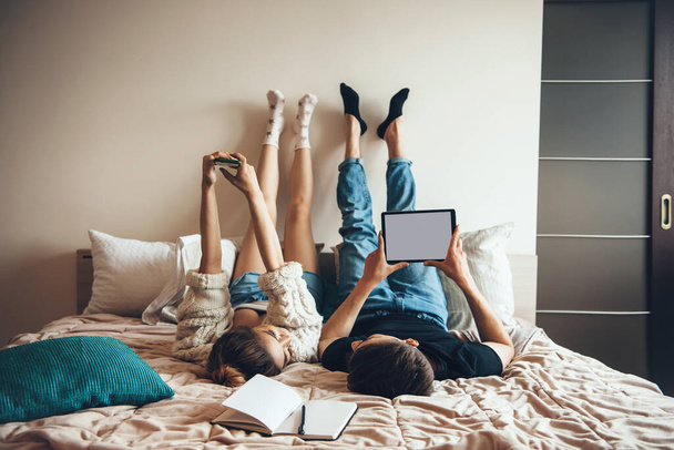 Zadní pohled fotografie bělošského páru ležícího v posteli s moderními technologiemi a usmívajícího se na sebe pózujícího u knihy - Fotografie, Obrázek