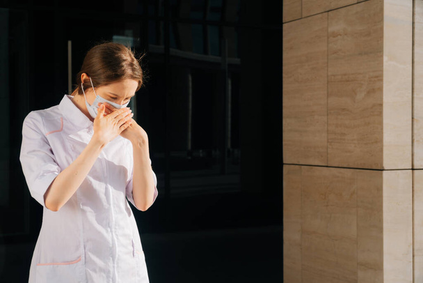 Caucasian young girl in a protective mask in a robe sneezes in the street. Female doctor experiences cough in front of hospital - Foto, Imagem