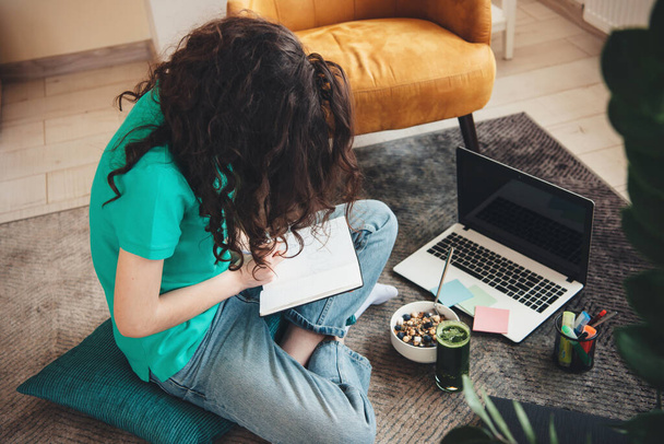 Oberes Foto einer kaukasischen Studentin, die Hausaufgaben auf dem Boden macht und einen Laptop benutzt, während sie Müsli mit Gemüsesaft isst - Foto, Bild