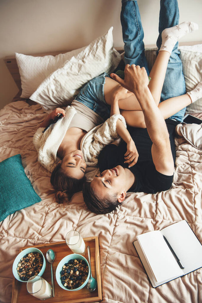 Horní pohled fotografie bělošského páru ležícího v posteli a objímajícího se před konzumací cereálií s mlékem - Fotografie, Obrázek