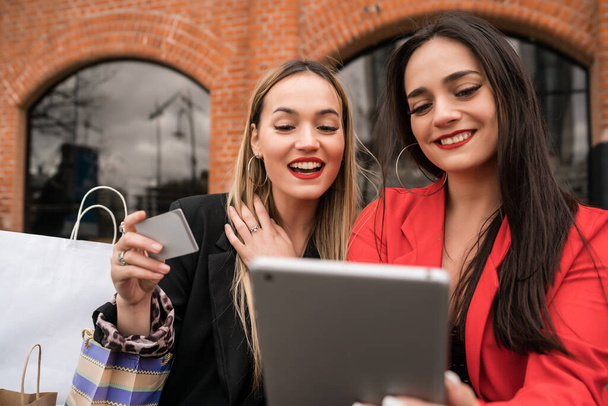 Portrait of two young friends shopping online with credit card and digital tablet while sitting outdoors. Friendship and lifestyle concept. - Foto, Imagen
