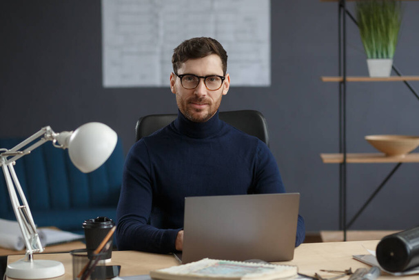 Architect working in office with laptop. Business portrait of handsome bearded man wearing eyeglasses sitting at workplace. Confident businessman became successful. Business concept - Foto, Imagen