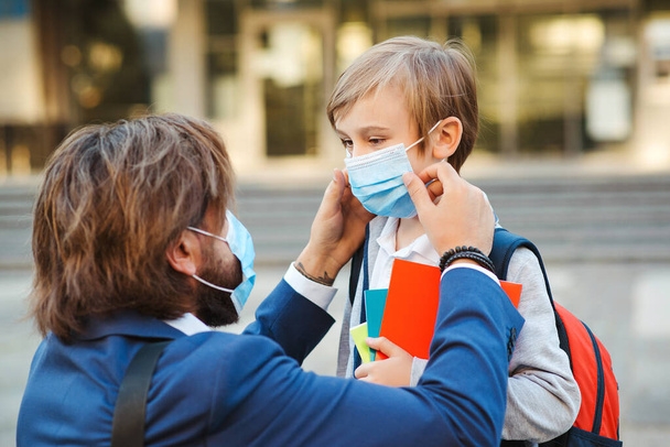 Father helping son and putting on face protective mask. Little boy going to school during coronavirus pandemic. Businessman father and son going to school. Schoolboy is ready go to school. - Foto, imagen