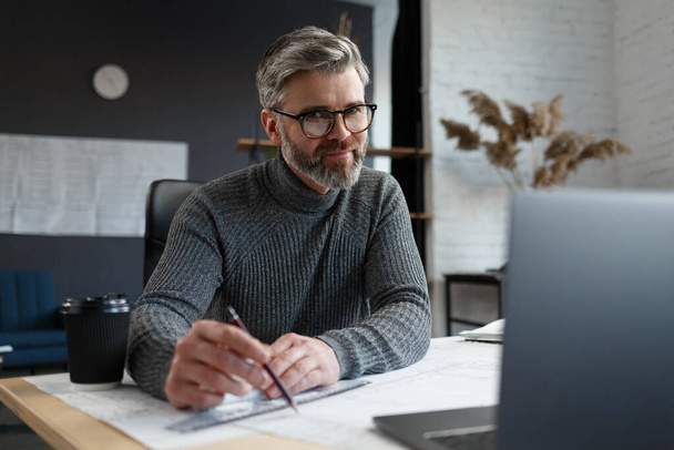 Interior designer working in office with blueprints.Engineer inspect architectural plan, sketching a construction project.Portrait of handsome bearded man at workplace. Business construction concept - Foto, Bild