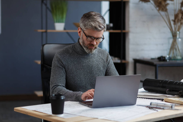 Designer working in office with laptop. Architect thinks over architectural plan, searching new ideas for construction project. Portrait of handsome bearded man sitting at workplace. Business concept - Fotografie, Obrázek