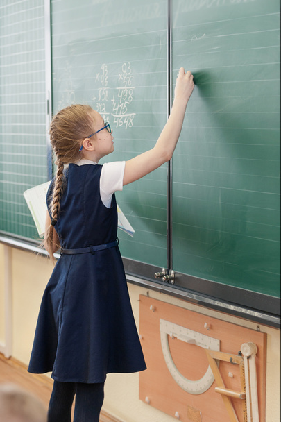 Dívka s culíkem v brýlích v modré uniformě na školní tabuli píše křídou s učebnicí v ruce - Fotografie, Obrázek