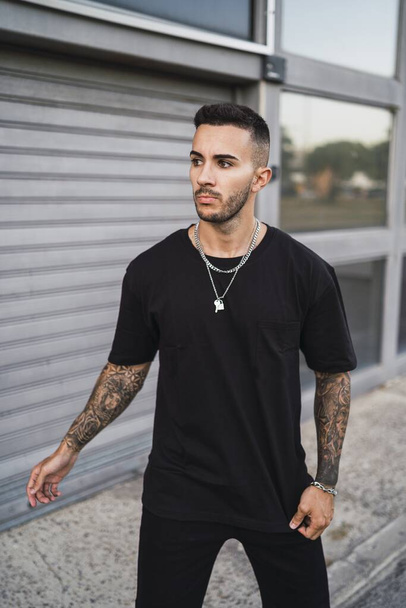 A vertical shot of a young European tattooed hip male in a black sporty outfit and a metal chain standing in front of a garage - Foto, Bild