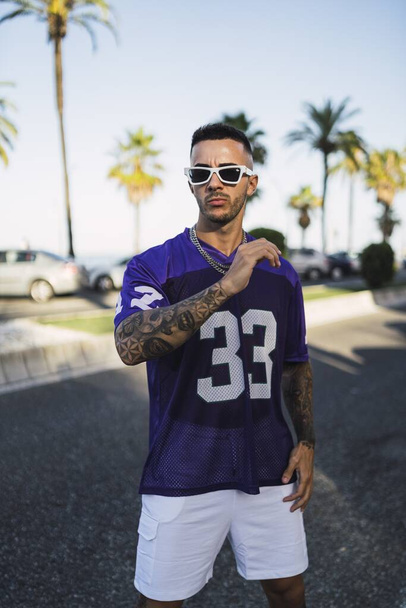 A vertical shot of a young European tattooed hip male in a purple and white sporty outfit with sunglasses posing in the street - Φωτογραφία, εικόνα
