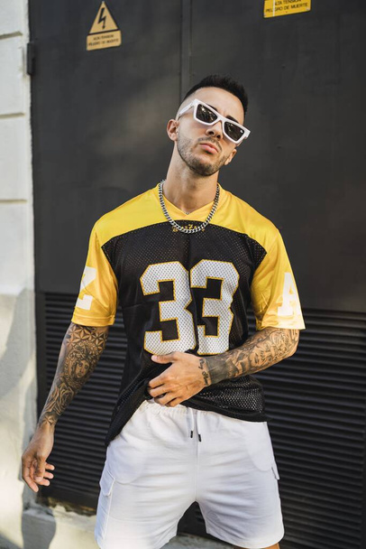 A vertical shot of a young European tattooed hip male in a yellow and black sporty outfit, sunglasses, and metal chain posing in front of a garage - Φωτογραφία, εικόνα