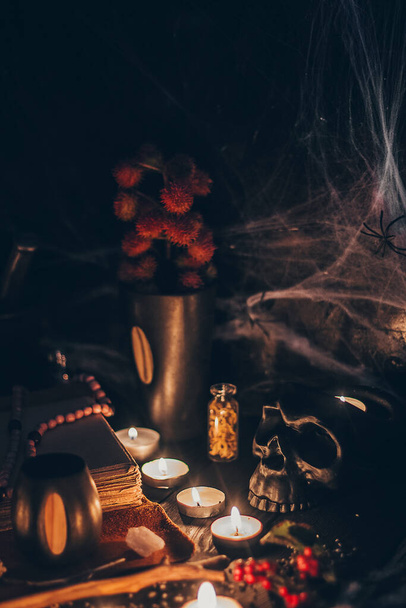 A ritual halloween witchcraft scene with candles, herbs, spider web, vintage bottles on the rustic background with a scary skull face and antique book - Photo, Image