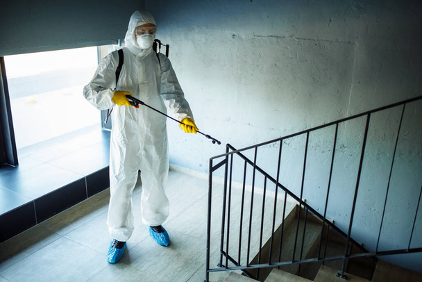 Sanierende Arbeiter säubern das Treppenhaus des Einkaufszentrums mit einem Antiseptikum, um eine Ausbreitung von Covid-19 zu verhindern. Ein Mann im Desinfektionsanzug besprüht Treppen. Gesundheitswesen, Quarantäne und Hygienekonzept - Foto, Bild