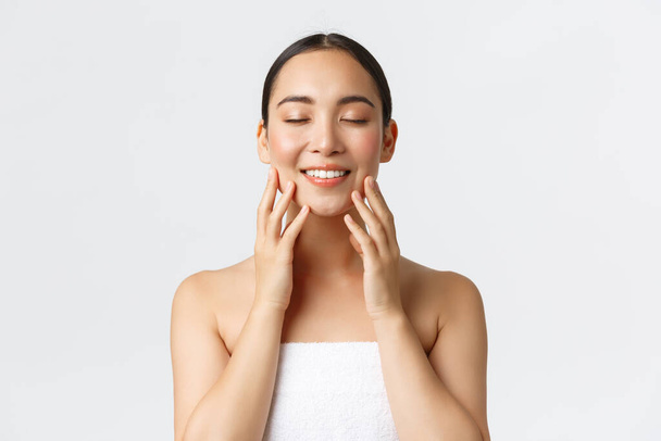 Tender smiling beautiful asian girl attend spa salon, standing in towel, close eyse and touching soft clean skin, satisfied with cream or cleansing gel results, standing white background - Photo, Image