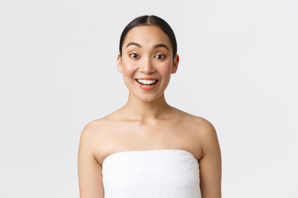 Opgewonden en vrolijk lachende Aziatische vrouw in handdoek kijken geïntrigeerd op de camera, zeggen wow, horen over spa salon kortingen, speciale aanbieding op badkamer en huidverzorgingsproducten, witte achtergrond - Foto, afbeelding