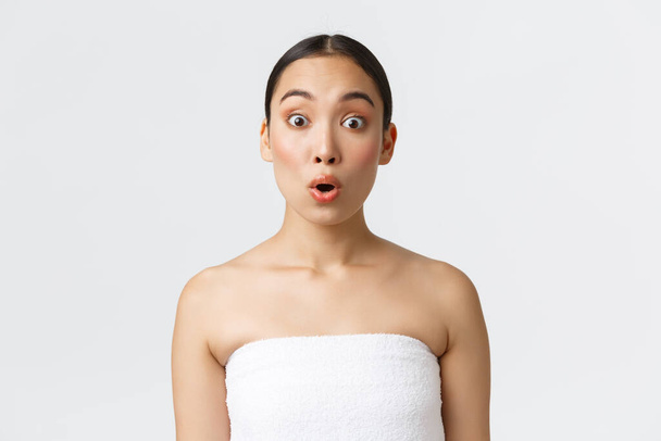 Εντυπωσιασμένη και περίεργη Ασιάτισσα ακούει ενδιαφέρουσα promo προσφορά, στέκεται σε πετσέτα πάνω από το λευκό φόντο, διαφήμιση του σπα σαλόνι, προϊόντα ομορφιάς, προϊόντα περιποίησης του δέρματος, λευκό φόντο - Φωτογραφία, εικόνα