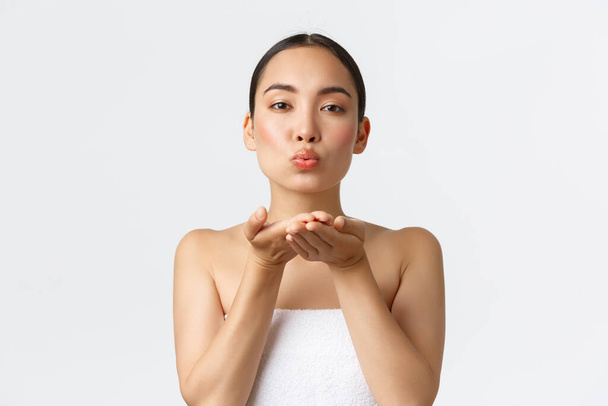 Szexi női ázsiai lány törölközőben küld levegő csók gyengéden a kamera, promo kínál bőrápoló termékek, sampon vagy arc tisztító. Nő áll a fürdőszobában, és fúj mwah - Fotó, kép