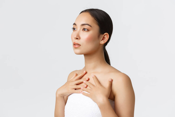 Profil kobiecej zmysłowej młodej Azjatki w białym ręczniku patrzącej w lewy górny róg i dotykającej jej ciała, promocja kosmetyków, produktów do pielęgnacji skóry lub kremów do twarzy, białe tło - Zdjęcie, obraz