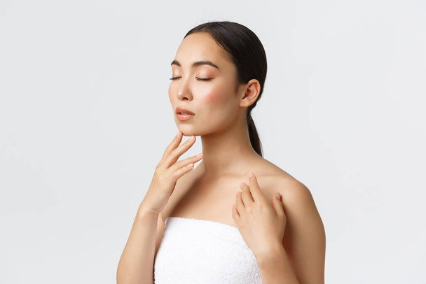 Feminine and tender young asian woman in towel close eyes, gently touching clean perfect skin, applying skincare product, visit spa or beauty salon, standing white background - Photo, Image