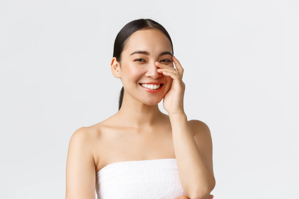 Concepto de salón de belleza, cosmetología y spa. Primer plano de hermosa mujer asiática en toalla de baño riendo y sonriendo dientes blancos, suavemente tocando la cara, la piel limpia y el concepto de cuidado de la piel
 - Foto, imagen