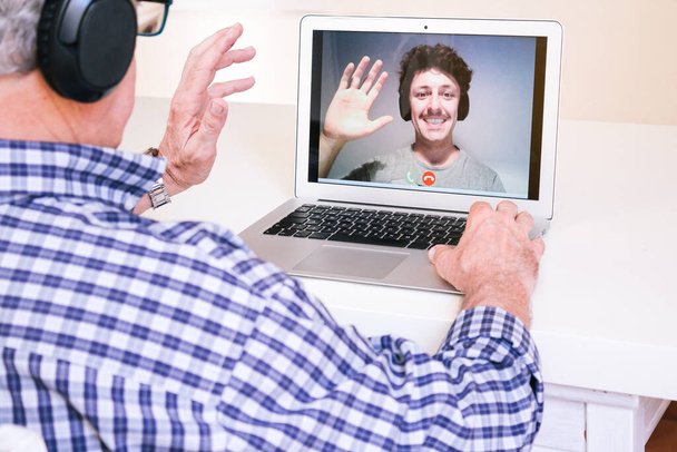 Kulaklıklı yaşlı bir adam laptopunda oğluyla video görüşmesi yaparken birbirini mutlu bir şekilde selamlıyor.. - Fotoğraf, Görsel