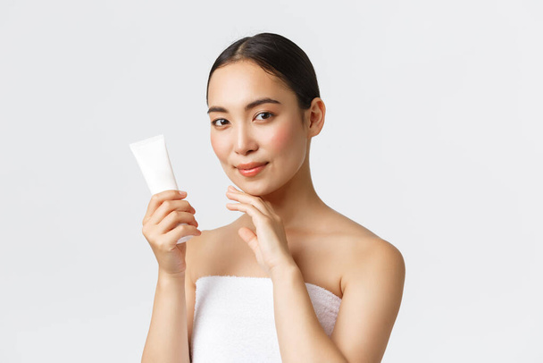 Beauty, personal care, spa salon and skincare concept. Young beautiful asian woman in bath towel touching face and showing facial cream, moisturizing treatment of skin, standing white background - Photo, Image
