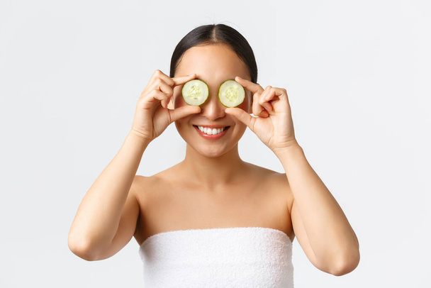 Beauty, personal care, spa salon and skincare concept. Close-up of beautiful smiling asian female in bath towel holding cucumber on eyes, promoting moisturzing, hydration cream or skin product - Fotó, kép
