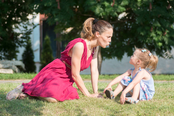 Мати в червоній сукні грає зі своєю милою дочкою на відкритому повітрі на траві, єдність і сім'я
. - Фото, зображення