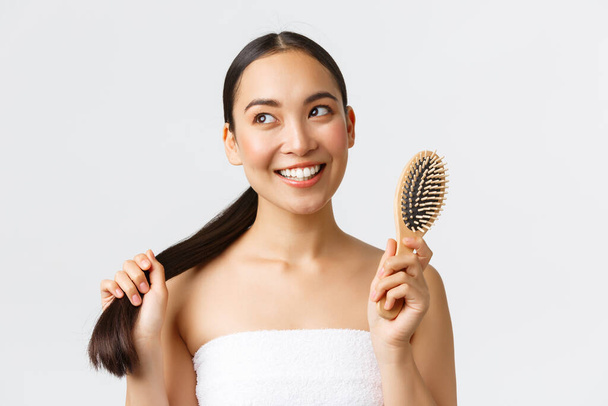 Beauty, hair loss products, shampoo and hair care concept. Dreamy beautiful asian girl in bath towel looking away with coquettish smile, brushing ponytail with brush over white background - Foto, Bild