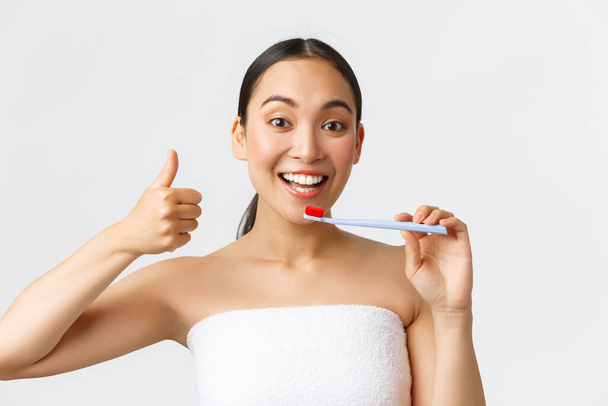 Beauty, personal care, and hygiene concept. Pleased attractive asian woman in bath towel trying new toothbrush, showing thumb-up great toothpaste for teeth whitening, white background - Valokuva, kuva